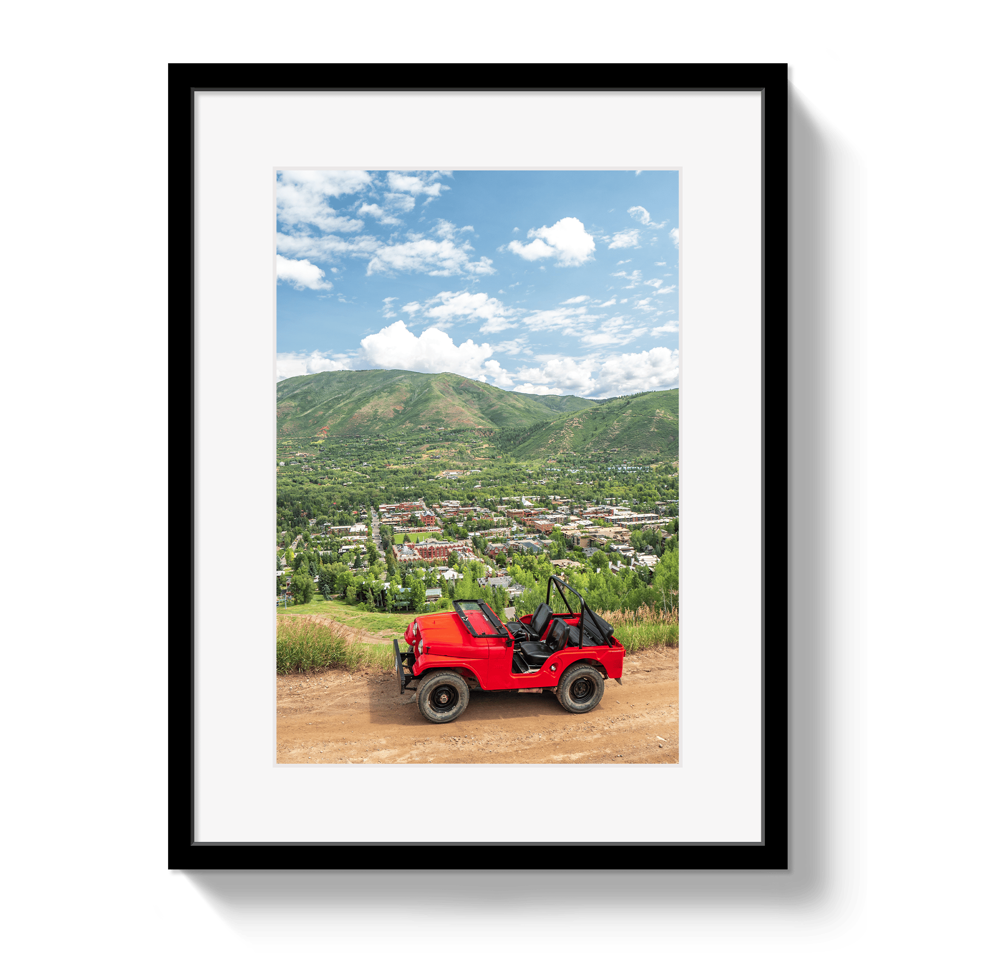 Framed photograph of a red jeep on a dirt road with mountains and town in the background.