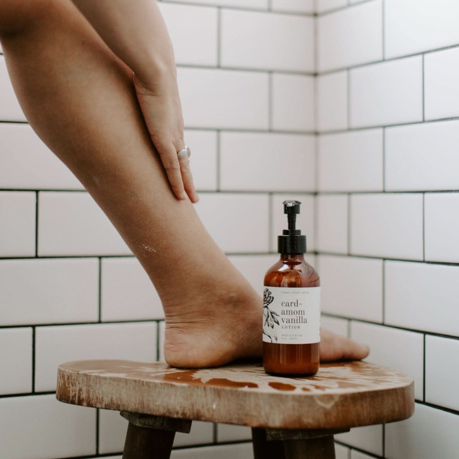 Person standing on a wooden stool with a bottle of body wash in a tiled bathroom.