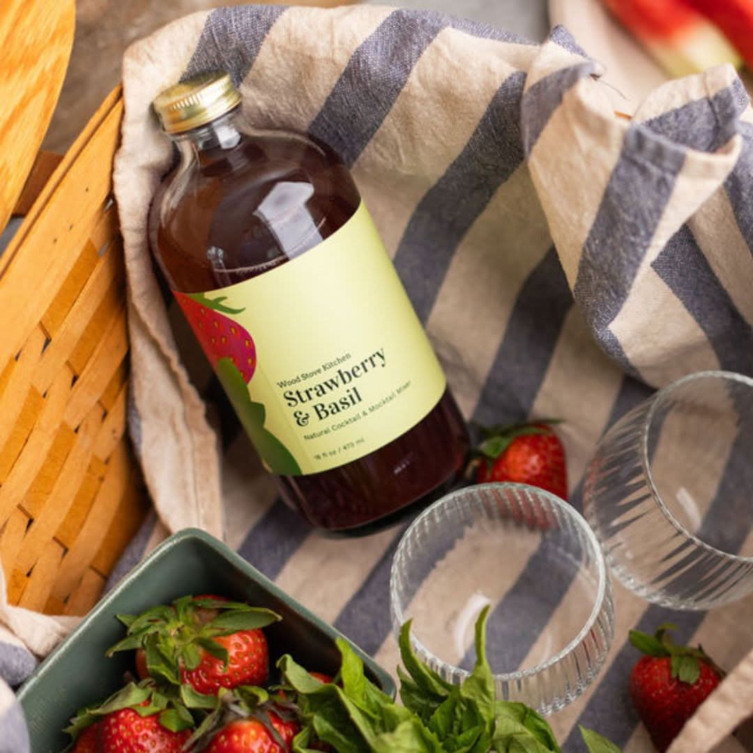 Bottle of strawberry basil syrup on a striped cloth with strawberries and a basket in the background