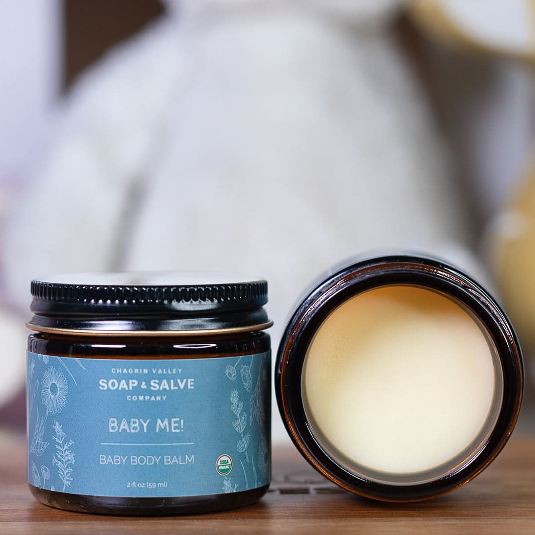 Baby body balm jar with open lid on a wooden surface, featuring the &