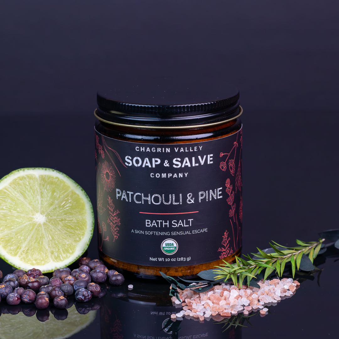 A jar of Chagrin Valley Soap & Salve Patchouli & Pine Bath Salt is displayed on a reflective dark surface with a lime half, blueberries, pink salt, and a sprig of pine. The product features a black label with white and red text, set against a dark background.