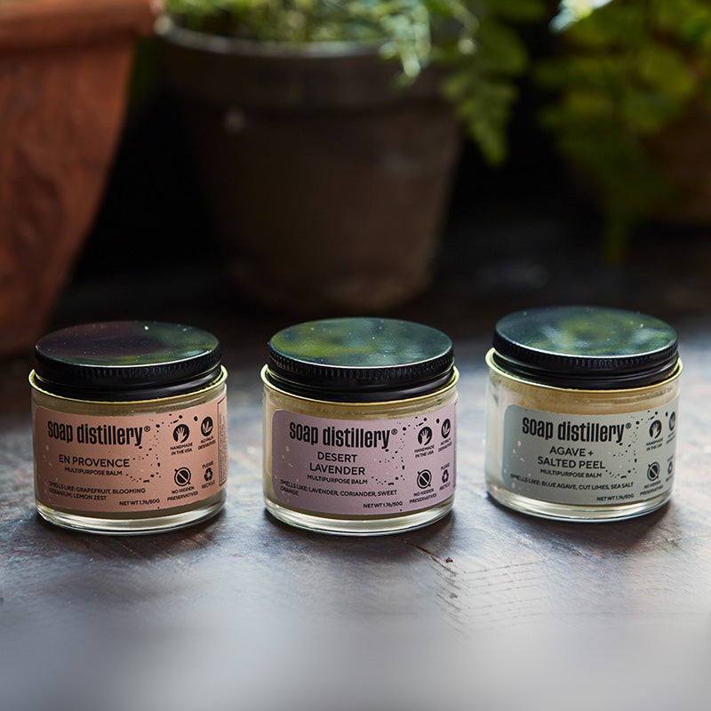 Three jars of Soap Distillery products on a wooden surface with plants in the background.