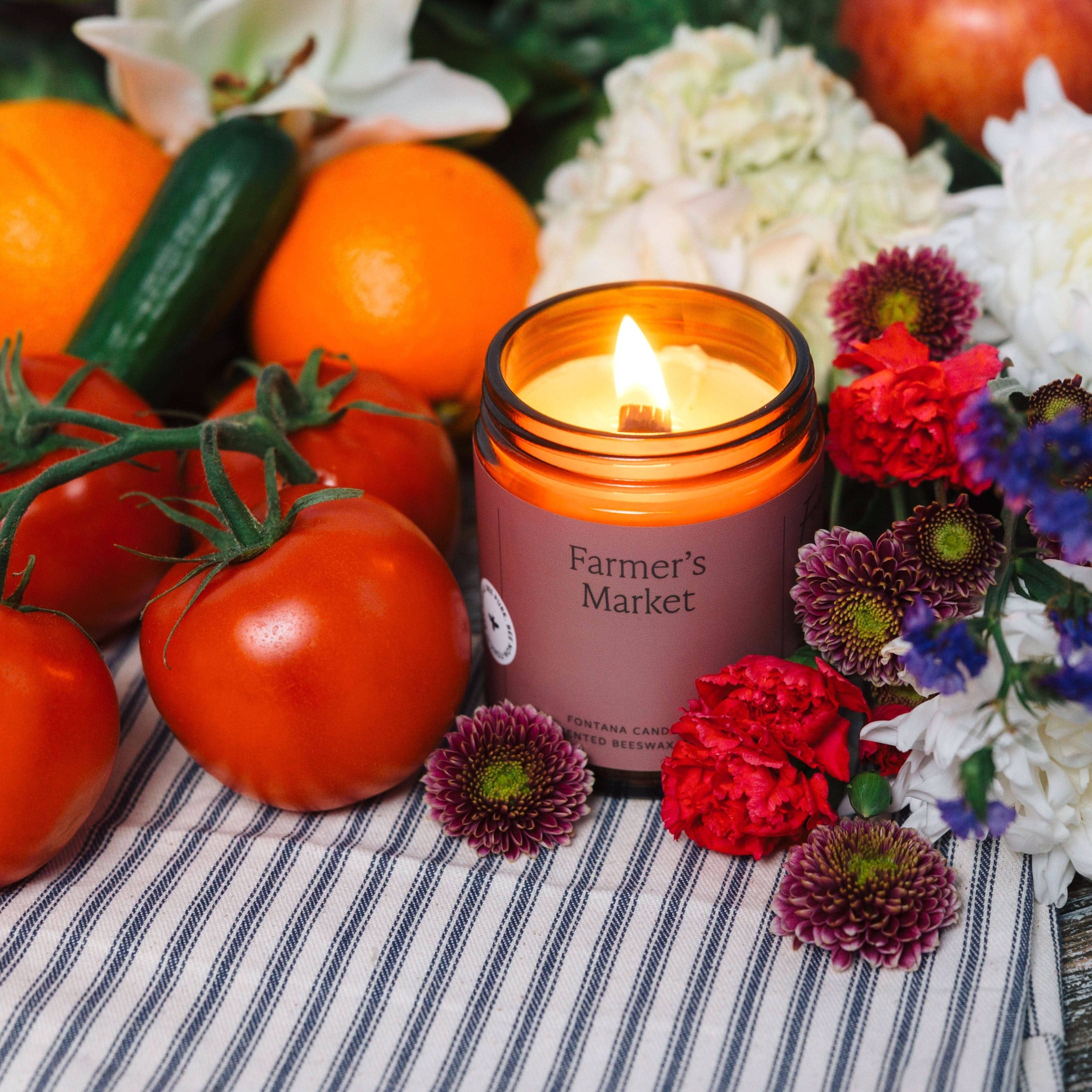 Candle labeled 'Farmer's Market' surrounded by tomatoes, oranges, and flowers on a striped fabric background