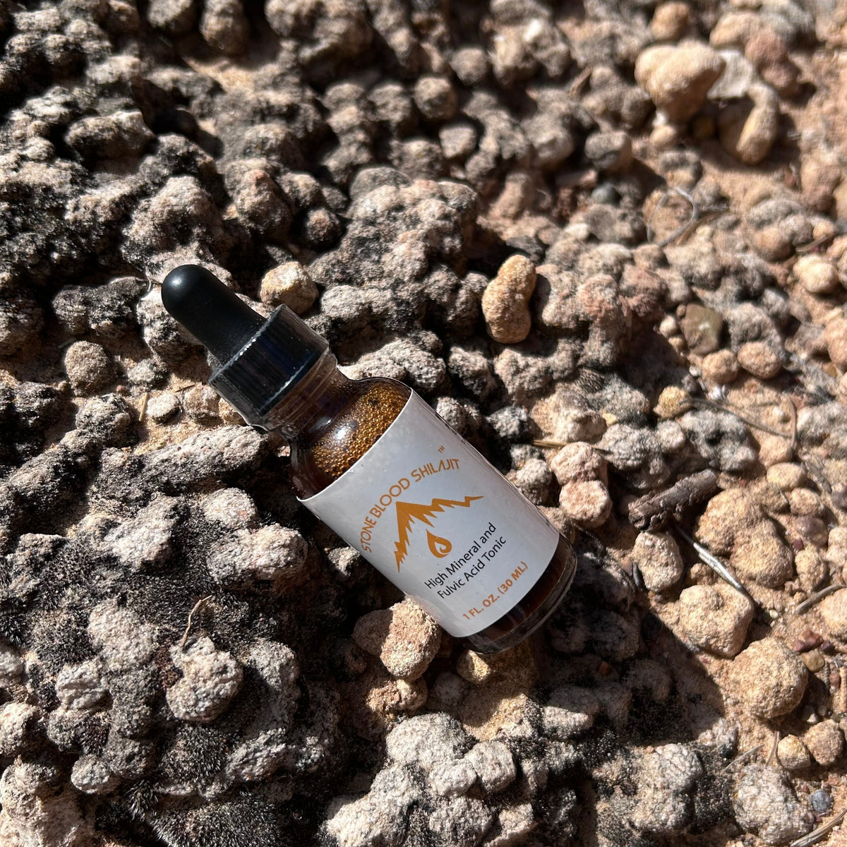 A small amber glass dropper bottle with a white label lies on rough, rocky, pebble-covered ground outdoors in bright sunlight.