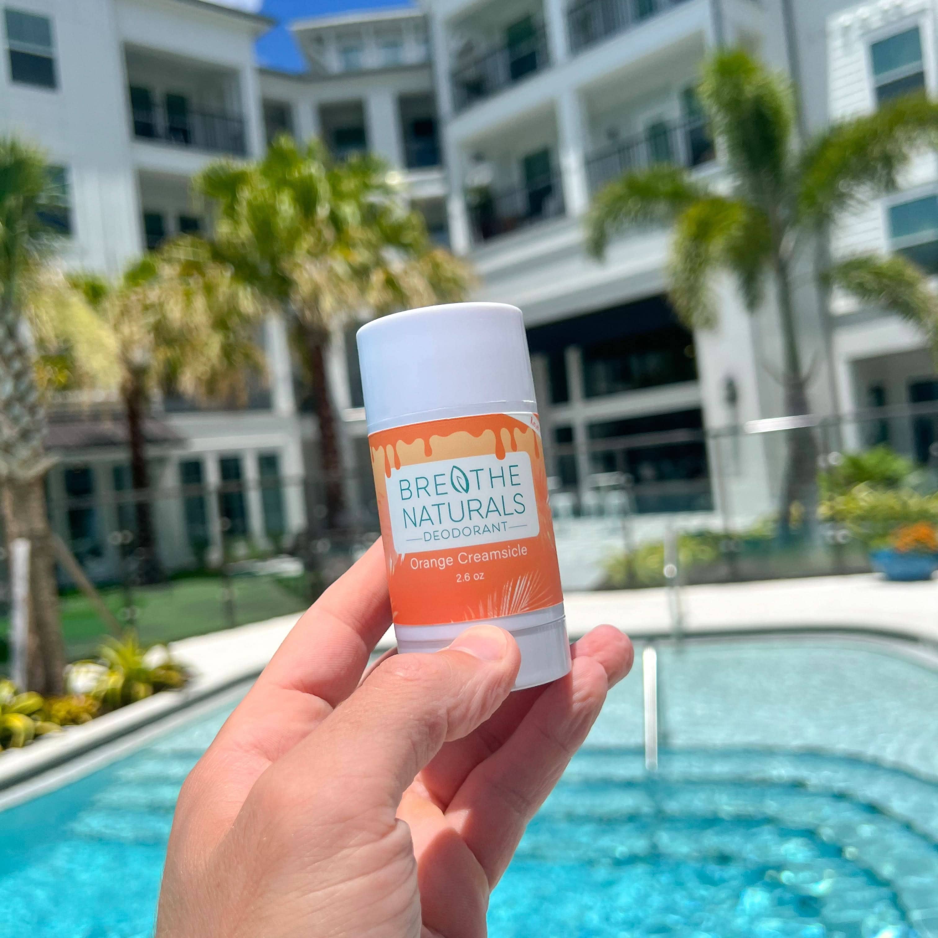 Hand holding a Breathe Naturals deodorant stick in front of a pool and building.