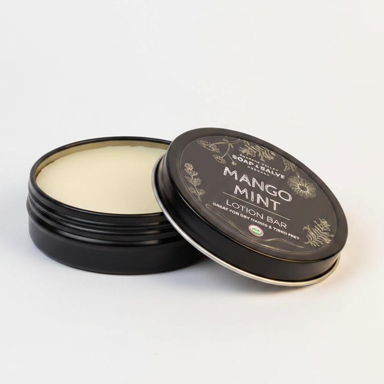 Mango Mint lotion bar in a black container on a dark surface with dried lemon and leaves.
