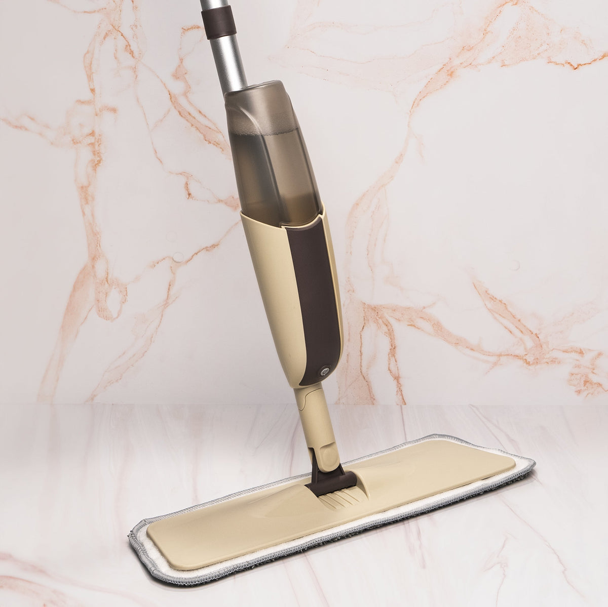 A steam mop with a beige and dark brown body, shown from a top-down angle as it cleans a light beige microfiber pad on a white marble floor against a matching marble wall.