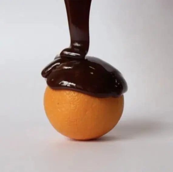 A bright orange fruit topped with glossy dark chocolate sauce, photographed against a clean light gray background.