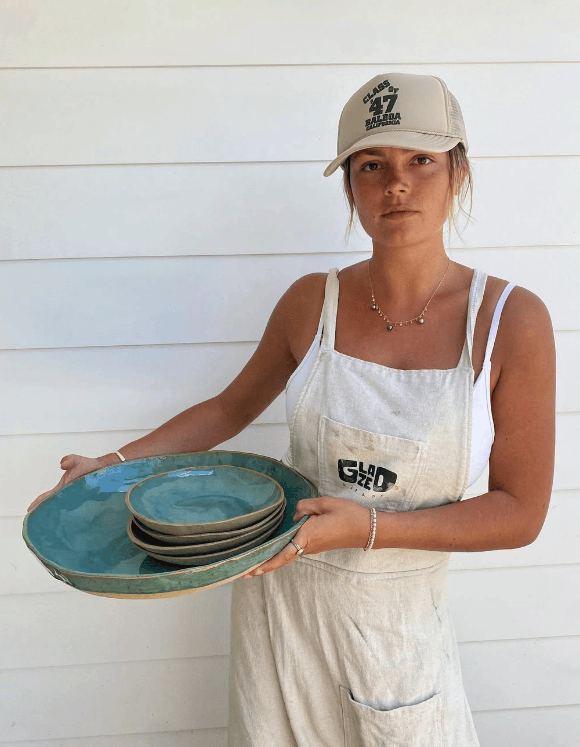Person holding a set of ceramic plates wearing a cap and apron against a white wall.