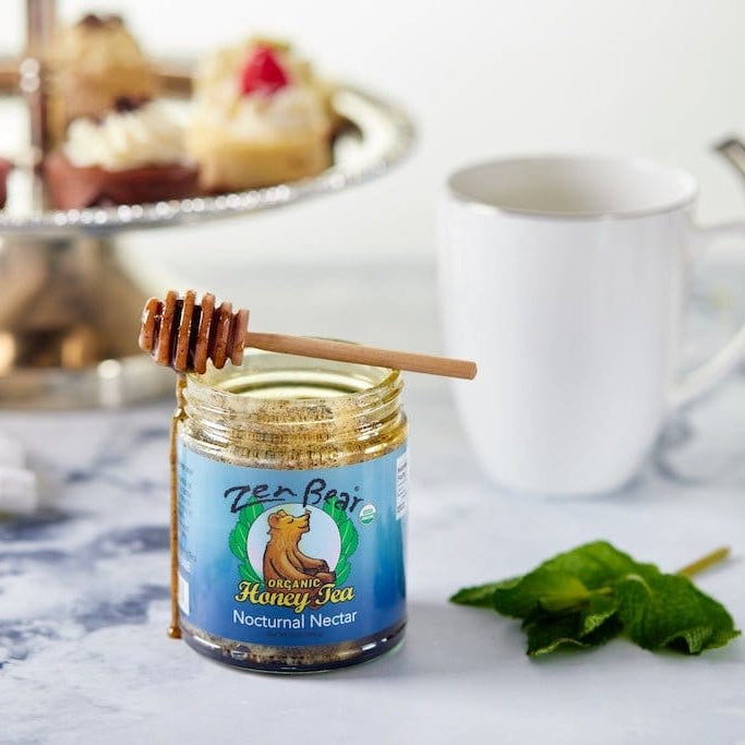 A jar of Zer Brax Honey Tea Nocturnal Nectar with a wooden honey dipper resting on the lid, set on a marble surface next to a white mug, a silver teapot, and green leaves, with cupcakes blurred in the background.