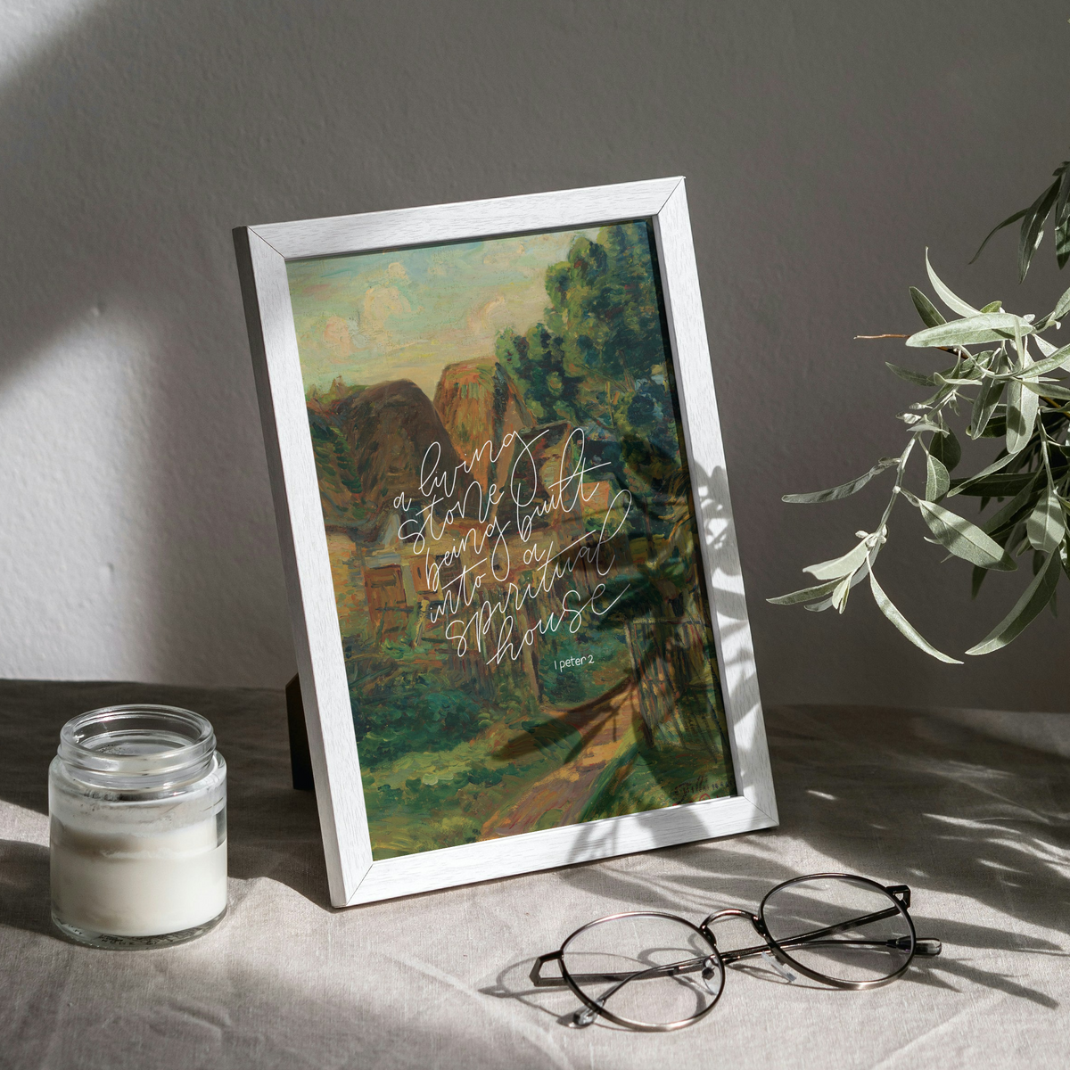 A framed landscape art print with a white frame, leaning on a draped fabric surface next to a candle jar, eyeglasses, and a vase with green leaves. The scene is lit by dramatic, diagonal natural sunlight casting shadows on a textured gray wall.