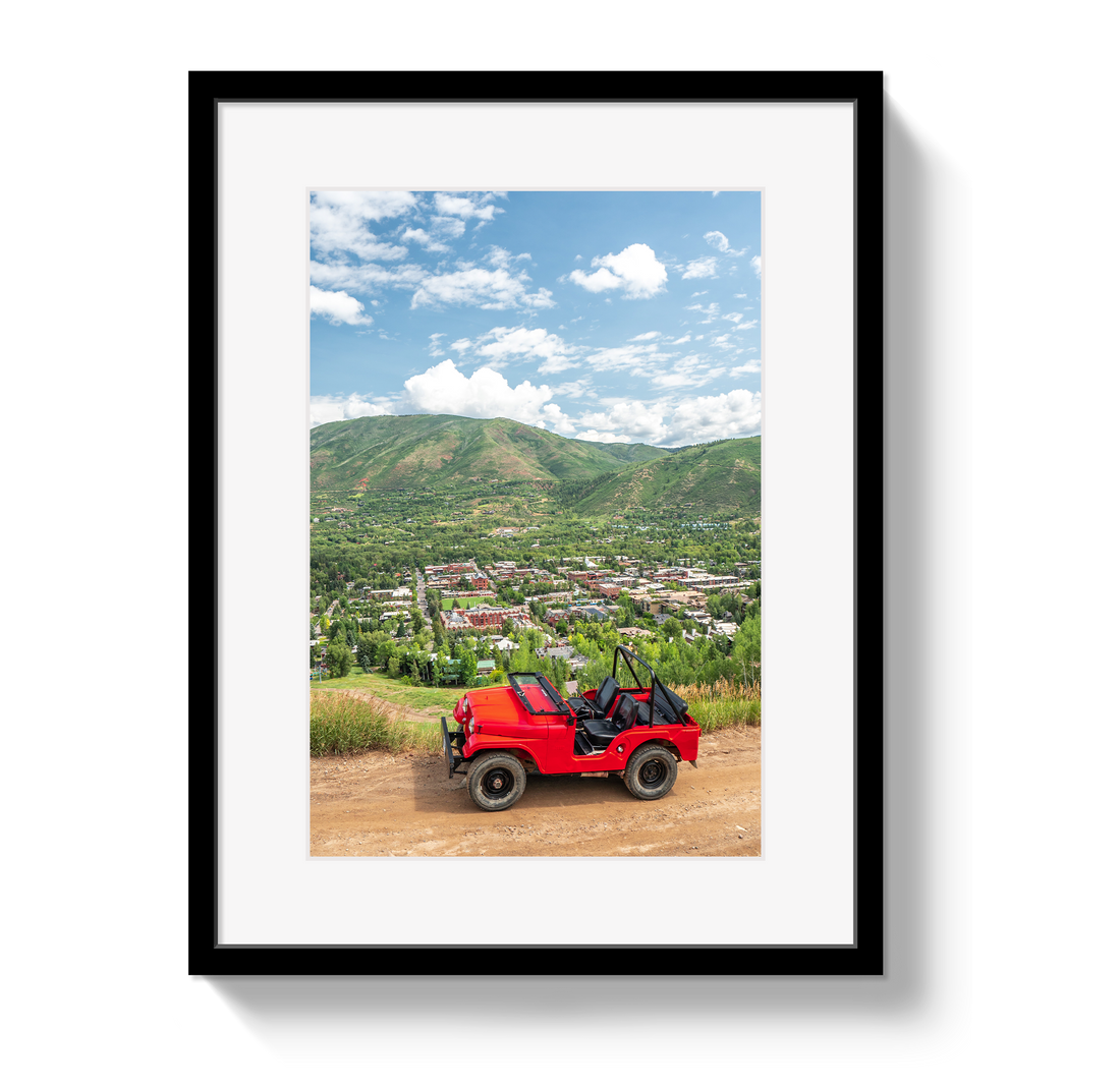 Framed photograph of a red jeep on a dirt road with mountains and town in the background.