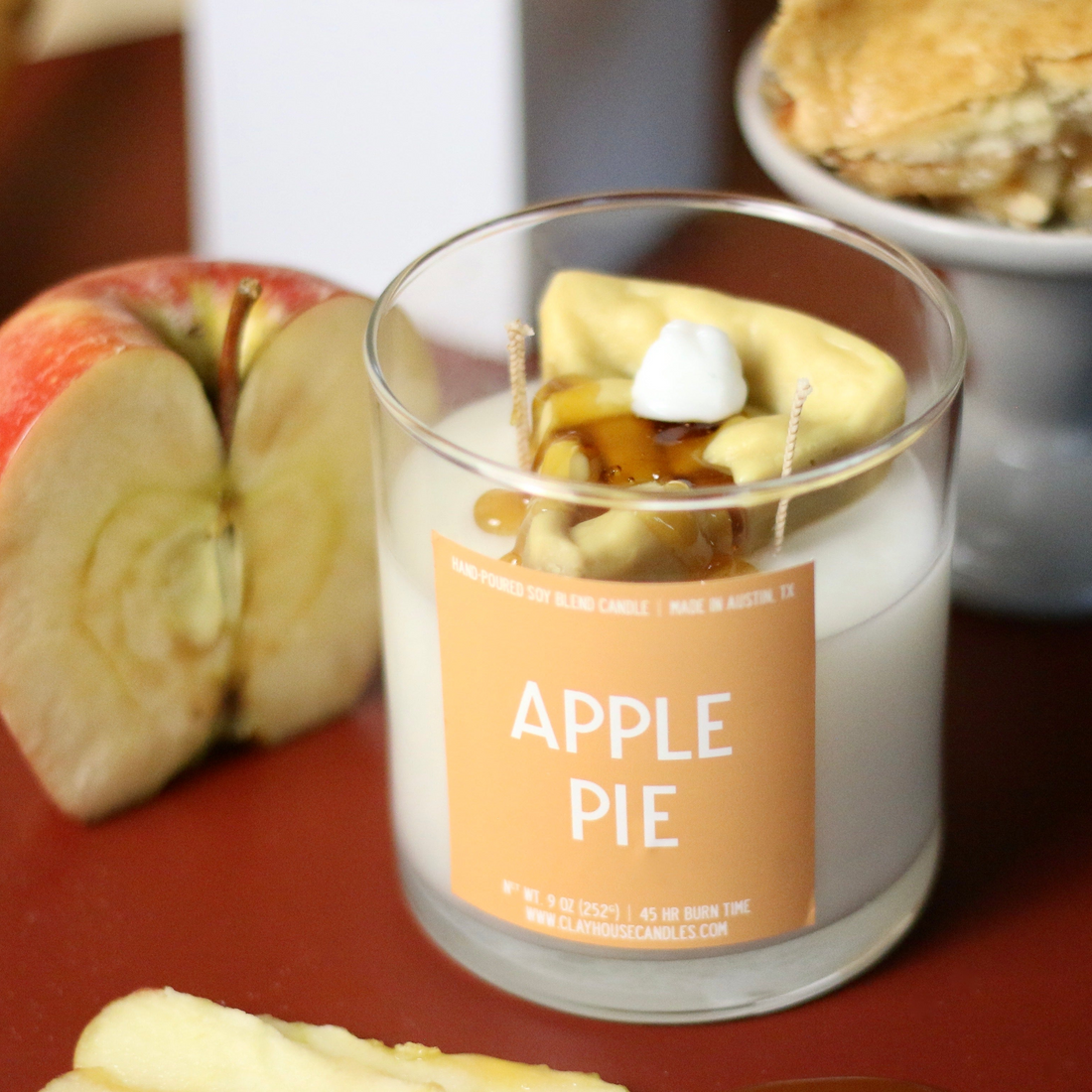 An Apple Pie scented candle in a clear glass jar with wax and decorative apple pieces on top, set on a dark red surface with sliced apples and a pastry in the background.