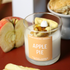 An Apple Pie scented candle in a clear glass jar with wax and decorative apple pieces on top, set on a dark red surface with sliced apples and a pastry in the background.
