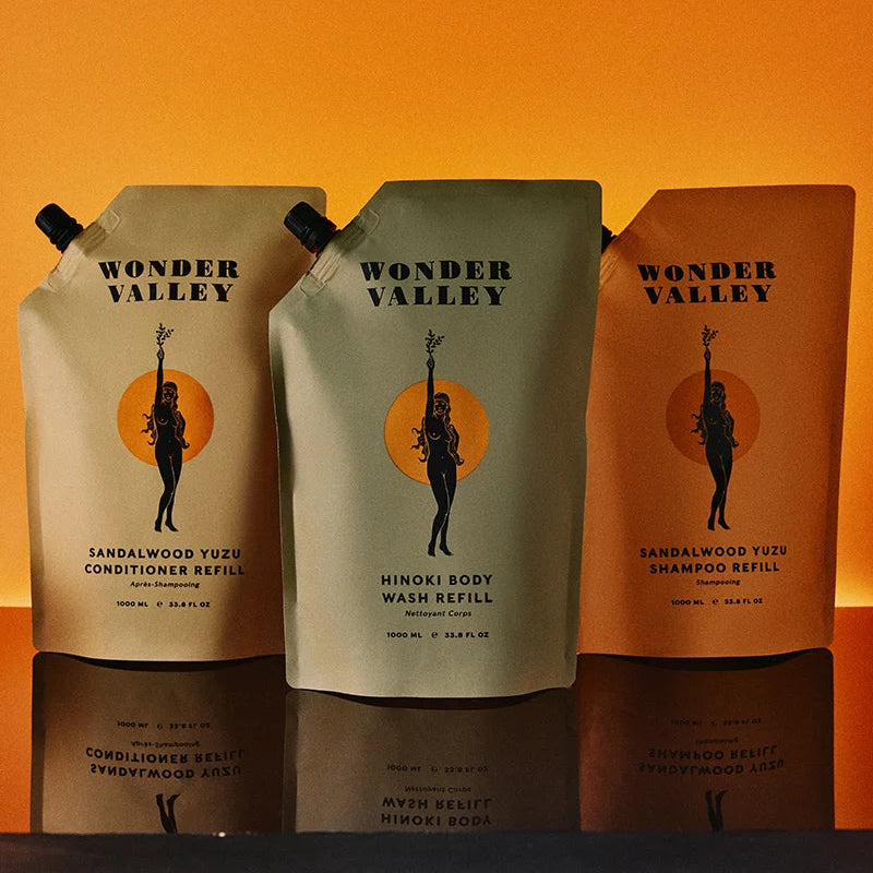 Three Wonder Valley refill pouches for hair care products are lined up on a reflective dark surface against a warm orange gradient background. The center pouch is a light gray refill with a black and orange logo, flanked by two orange-toned refill pouches.