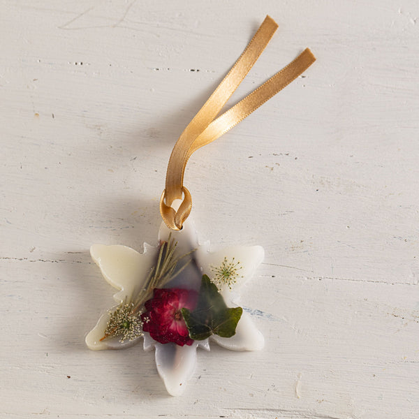 A white star-shaped resin ornament featuring a dried red rose, green leaves, and small wildflowers, suspended from a gold ribbon against a white wooden background.