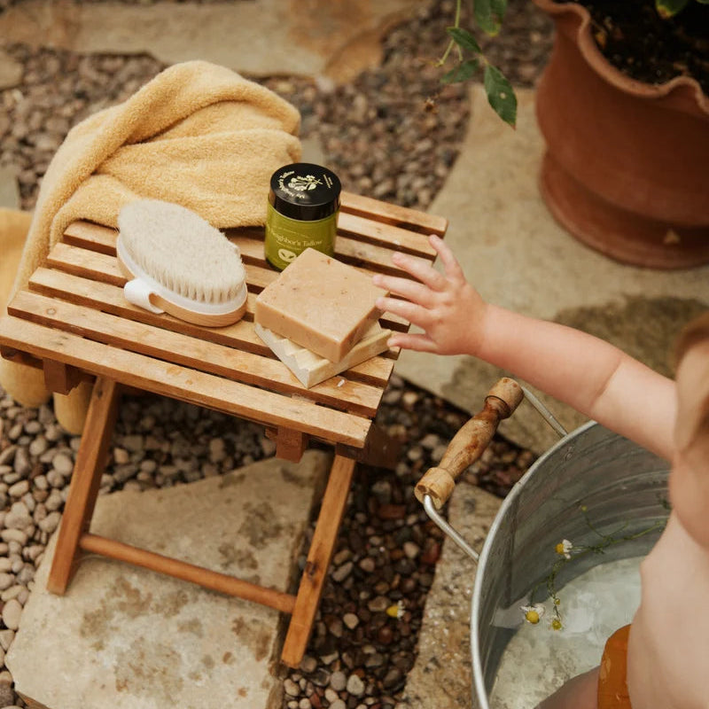 Person washing hands outdoors with a wooden stool, plant, and bucket.