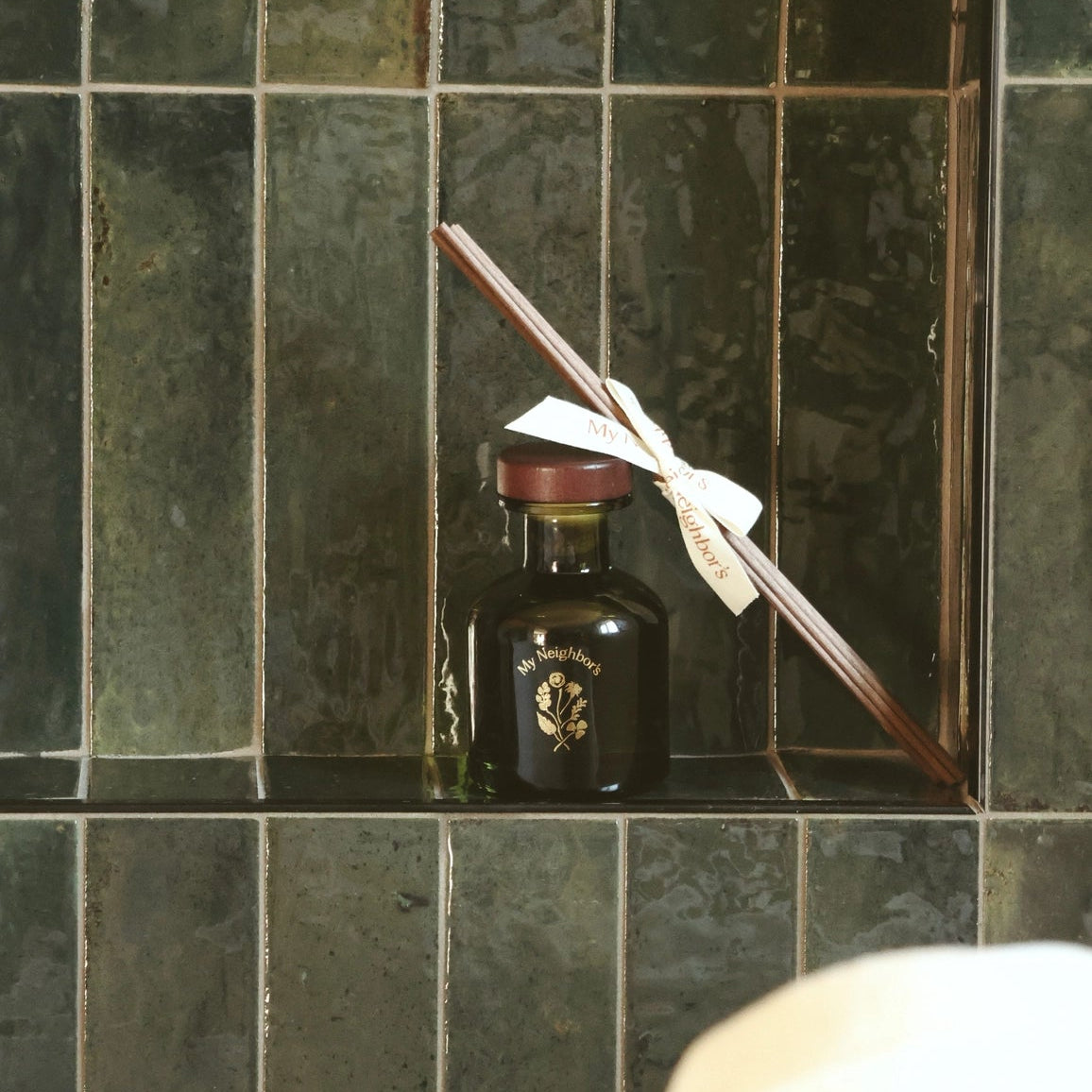 A dark glass bottle with a gold emblem and a white ribbon tied to a wooden stick, displayed on a tiled shelf.