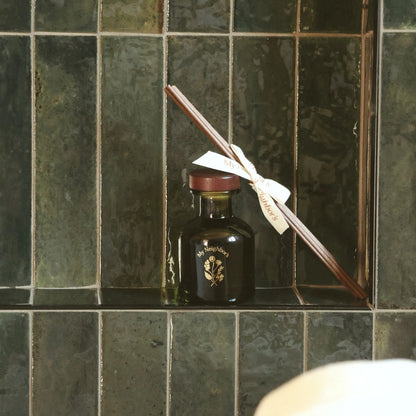 A dark glass bottle with a gold emblem and a white ribbon tied to a wooden stick, displayed on a tiled shelf.
