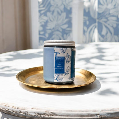 Candle jar with blue label on a gold tray in front of a window with leaf patterned curtains