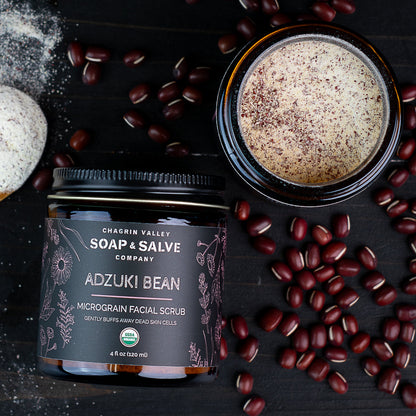 A jar of Chagrin Valley Soap &amp; Salve Company Adzuki Bean Micrograin Facial Scrub, shown next to an open jar displaying the creamy beige scrub with dark brown specks, surrounded by scattered adzuki beans on a dark wooden background.