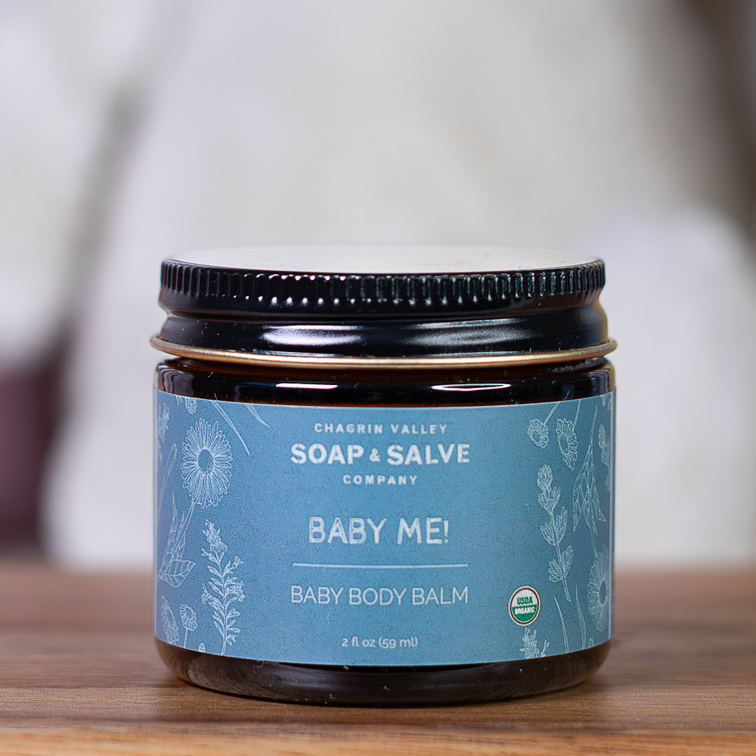 Jar of Chagrin Valley Soap &amp; Salve Company Baby Me! Baby Body Balm on a wooden surface with a blurred background.