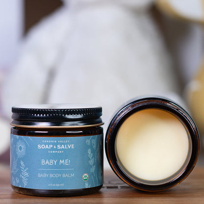 Baby body balm jar with open lid on a wooden surface, featuring the &