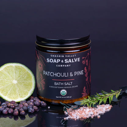 A jar of Chagrin Valley Soap &amp; Salve Patchouli &amp; Pine Bath Salt is displayed on a reflective dark surface with a lime half, blueberries, pink salt, and a sprig of pine. The product features a black label with white and red text, set against a dark background.