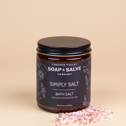 Chagrin Valley Soap &amp; Salve Company Simply Salt Bath Salt in an amber glass jar with a black lid and black label, shown on a dark reflective surface with pink salt crystals in front.