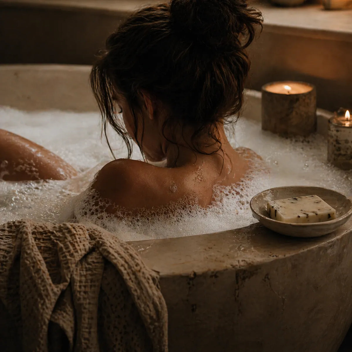 Person in a bathtub with bubbles, candles, and a bowl of soap.
