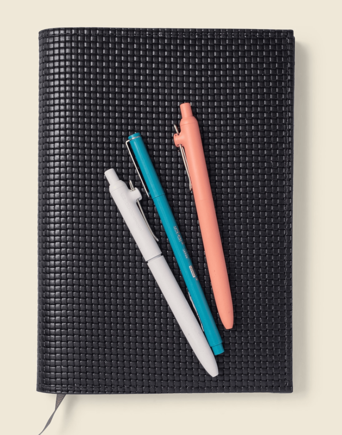 Black textured notebook with three pens on a light beige background