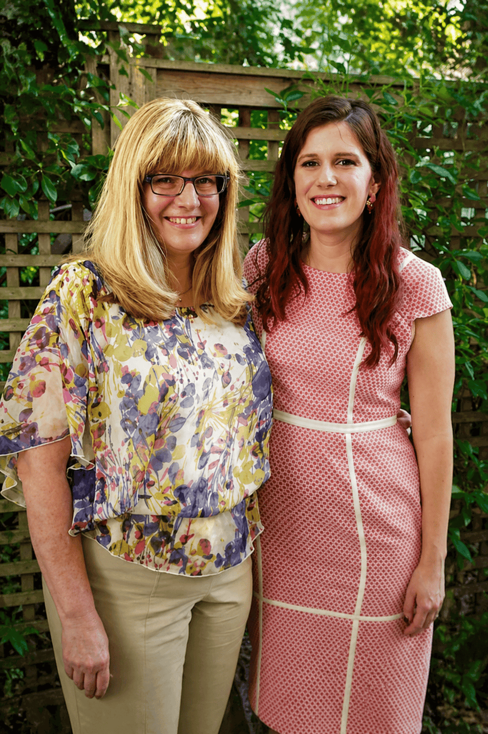 Two women standing together outdoors with a wooden fence and greenery in the background