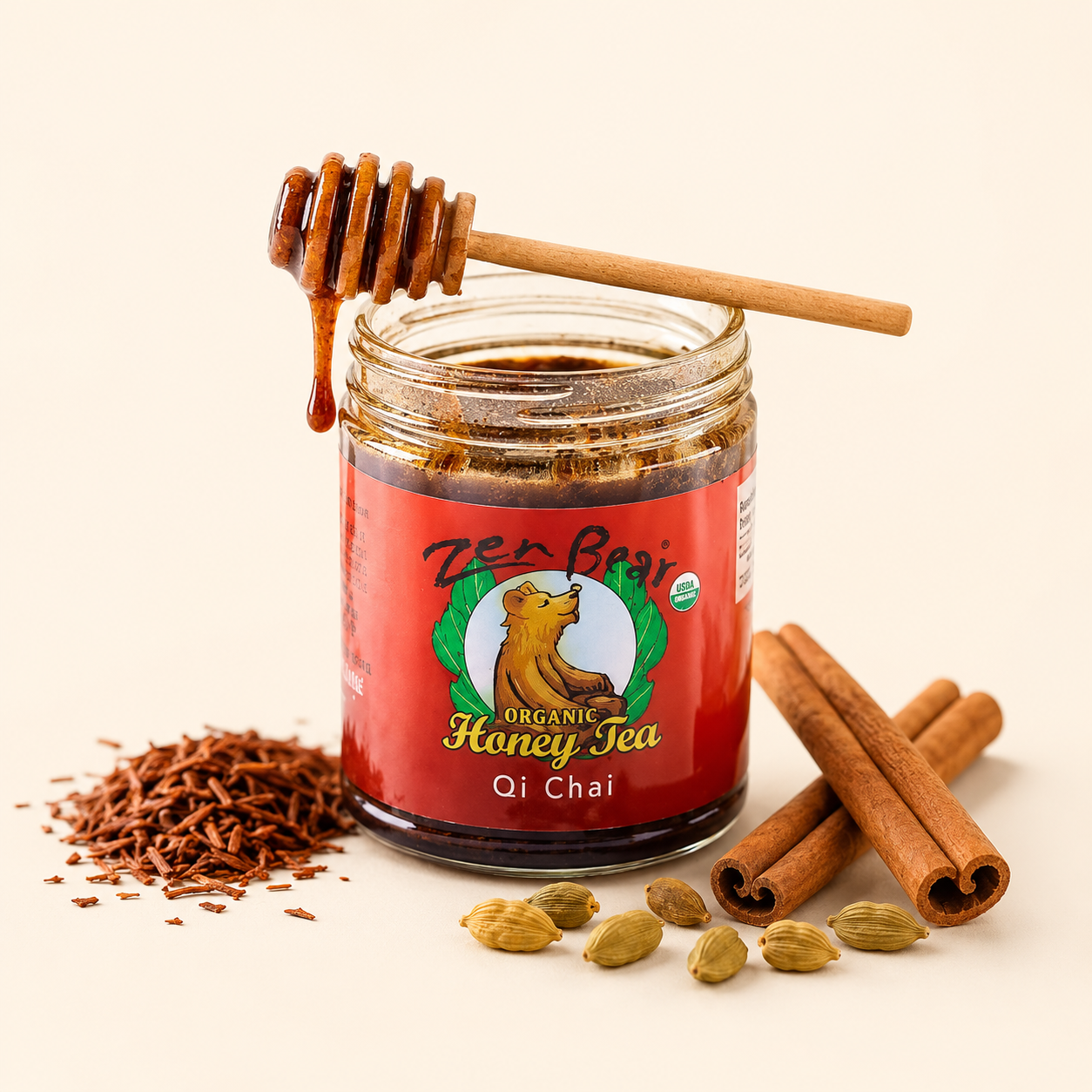 A glass jar of Zen Bear Organic Honey Tea Qi Chai with a red label and wooden honey dipper, styled with cinnamon sticks, cardamom pods, and loose brown powder on a light beige background.