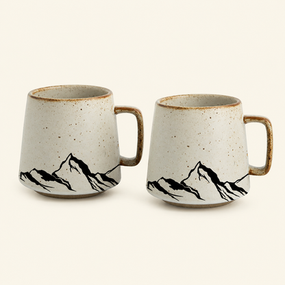 Two ceramic mugs with a speckled off-white glaze, brown rim, and black mountain line art, shown on a plain light beige background.