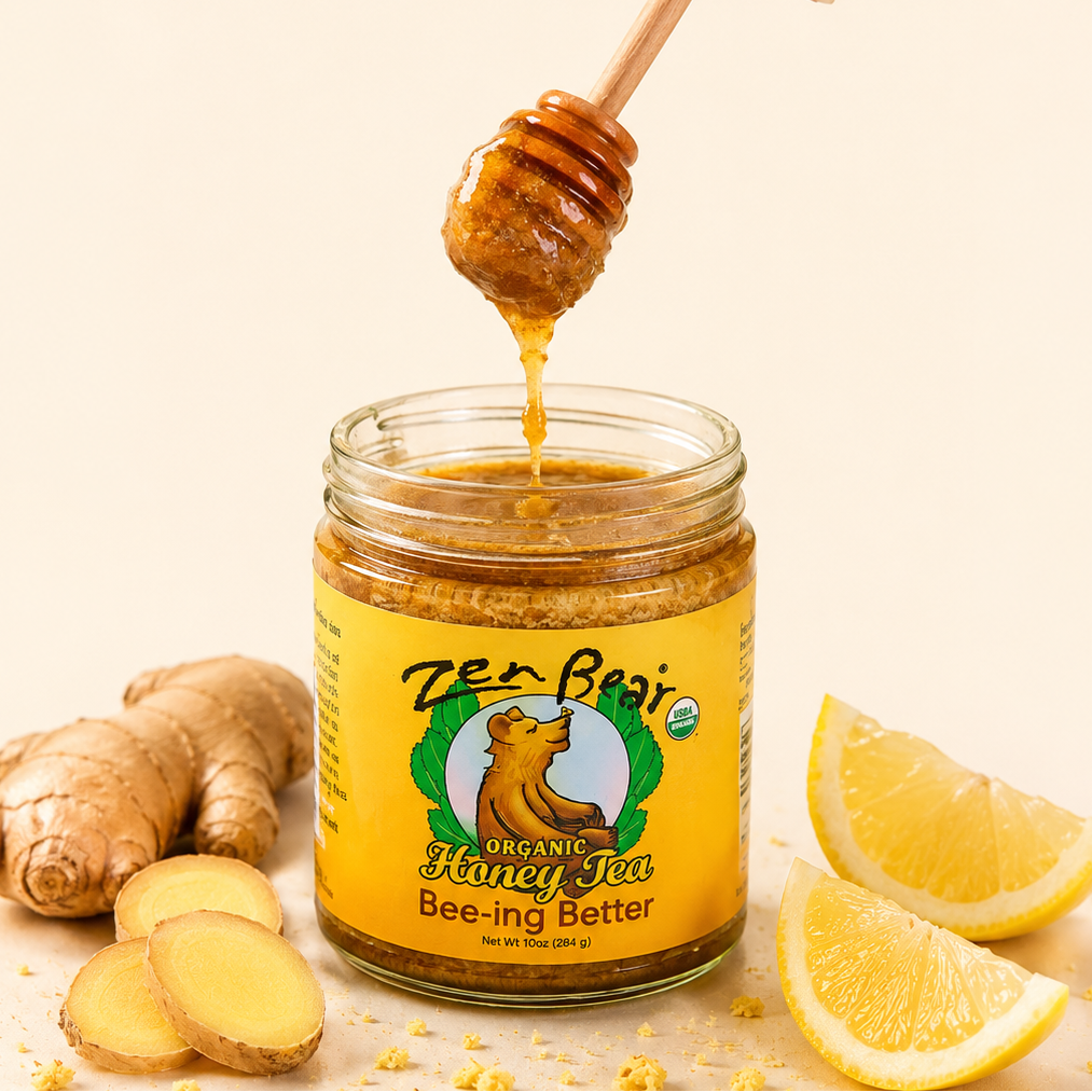A jar of Zen Beast Organic Mangosteen Bee-ing Better honey sits on a light beige surface with sliced ginger and lemon wedges around it. A hand holds a wooden honey dipper above the jar, with honey dripping into it.