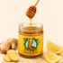 A jar of Zen Beast Organic Mangosteen Bee-ing Better honey sits on a light beige surface with sliced ginger and lemon wedges around it. A hand holds a wooden honey dipper above the jar, with honey dripping into it.