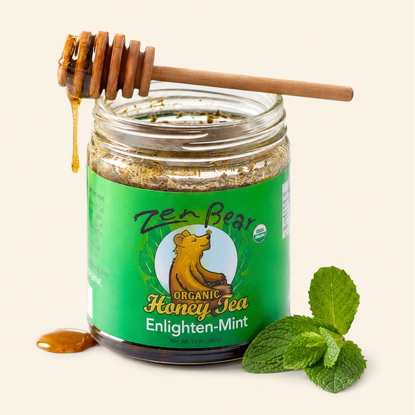 A jar of Zen Bear Organic Honey Tea Enlighten-Mint on a light cream background, with a wooden honey dipper dripping honey over the open jar and mint leaves beside it.