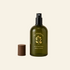 An olive green glass air and linen spray bottle with a black spray top and a separate brown cap, shown on a warm beige background. The bottle features a gold botanical logo and the brand name &