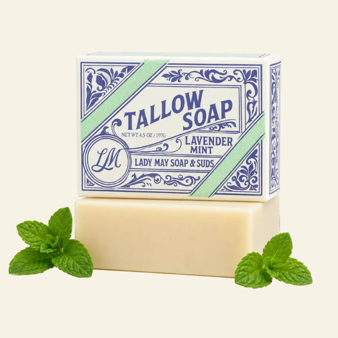 A bar of Tallow Soap from Lady May Soap &amp; Suds, specifically Lavender Mint, displayed in its blue and white patterned packaging box with a matching cream-colored bar of soap underneath, styled with fresh mint leaves and purple lavender sprigs on a white background.