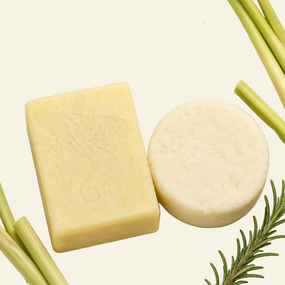 A shampoo and conditioner set made from natural-looking bars of soap, one rectangular pale yellow bar with embossed floral detailing and one round off-white bar, presented on a light cream background with rosemary sprigs and lemongrass stalks. Text at the top reads &