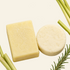 A shampoo and conditioner set made from natural-looking bars of soap, one rectangular pale yellow bar with embossed floral detailing and one round off-white bar, presented on a light cream background with rosemary sprigs and lemongrass stalks. Text at the top reads &