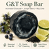 A G&T Soap Bar shown as a round dark charcoal bar with white marbled swirls, surrounded by blueberries, lime slices, and charcoal powder on a light beige background. Text at the top reads &