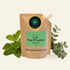 A stand-up pouch of Aster Earth MAG+IC Lotion Tingle shown on a light cream background with mint leaves, eucalyptus leaves, and crystals. The kraft paper pouch has a green label and a circular blue and yellow logo.