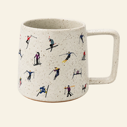 A speckled off-white ceramic mug with a handle, decorated with a repeating pattern of small skiers and snowboarders in various colorful outfits, set against a solid cream background.