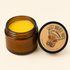 An open amber glass jar containing bright yellow balm or wax, shown next to its black lid with a kraft paper label. The label reads &