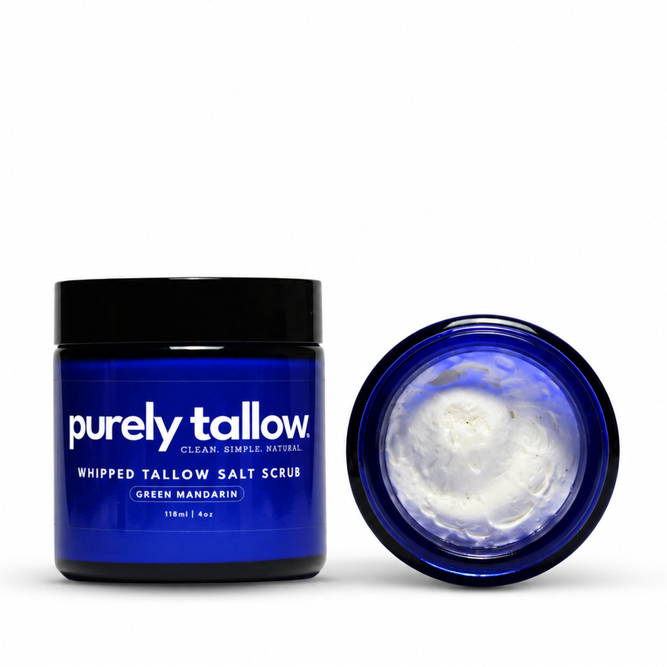 A dark blue jar of Purely Tallow Whipped Tallow Salt Scrub with a black lid, shown next to its open lid revealing white whipped cream inside, on a white background.