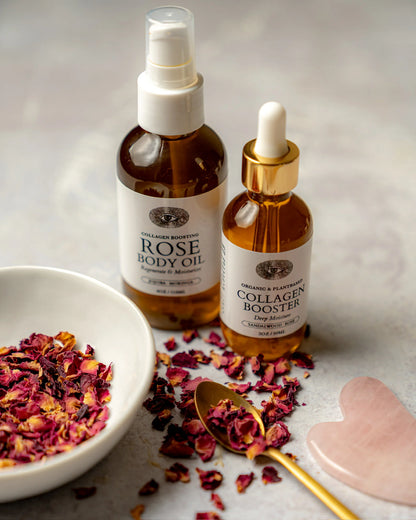 Two amber glass skincare bottles with white labels on a light stone surface, styled with dried rose petals in a white bowl, a gold spoon, and a pink stone. The larger bottle is Rose Body Oil, and the smaller dropper bottle is Collagen &amp; Flavour.
