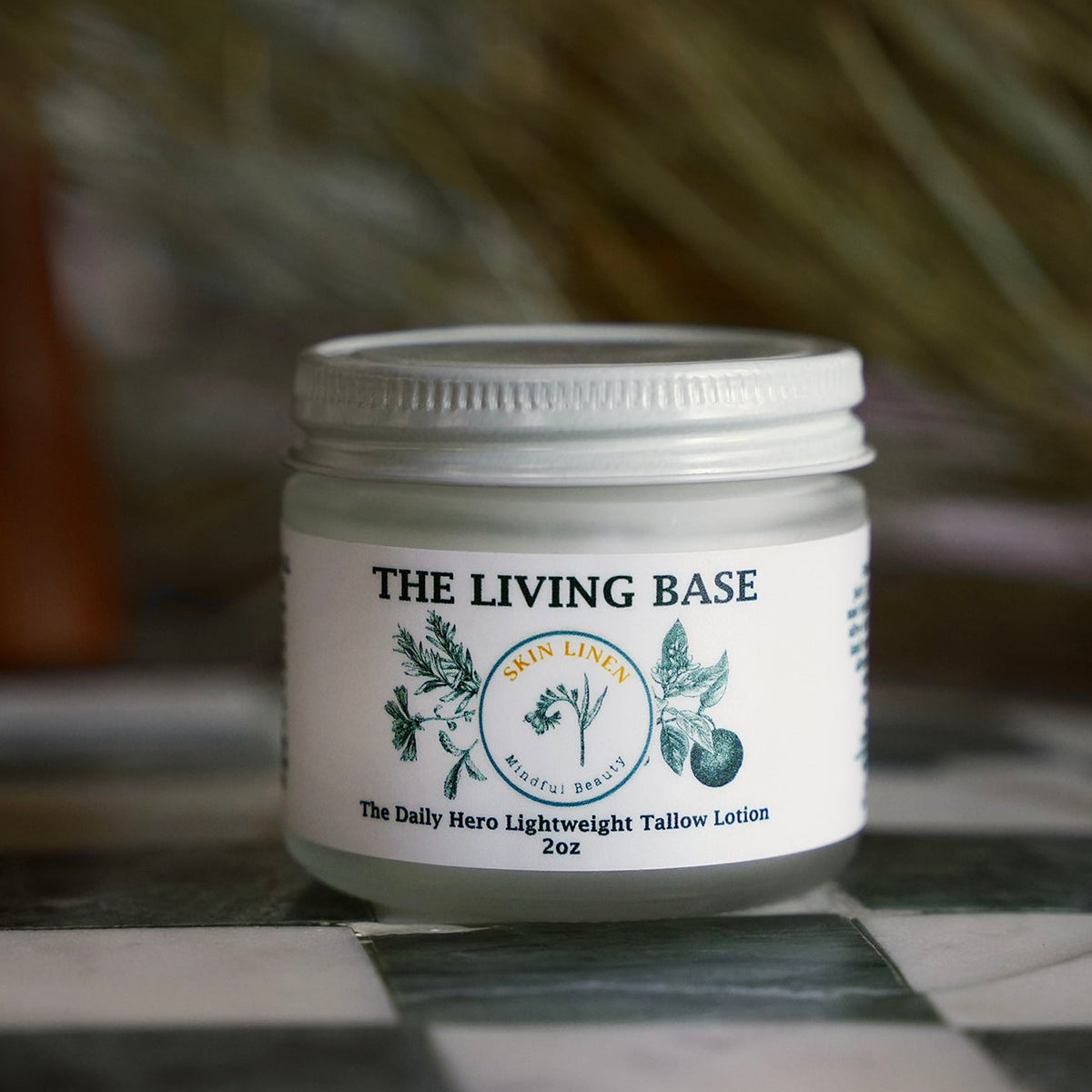 A small white jar of The Living Base Tallow Lotion sits on a black-and-white checkered surface with a blurred rustic background.
