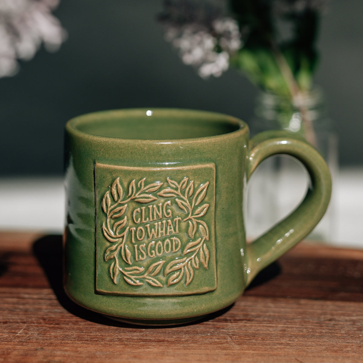 Green ceramic mug with embossed text on a wooden surface with flowers in the background