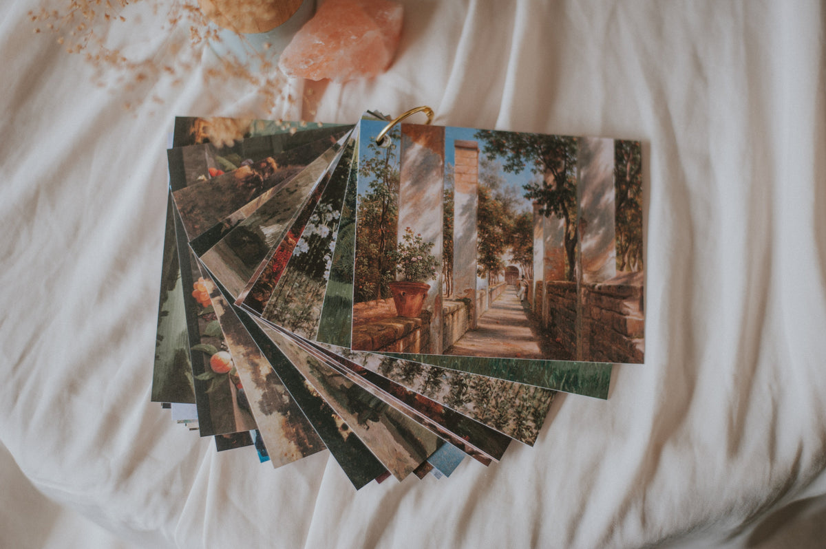 A fan of printed art cards or postcards featuring scenic landscape paintings, arranged on a white fabric surface with decorative dried flowers and a pink crystal nearby.